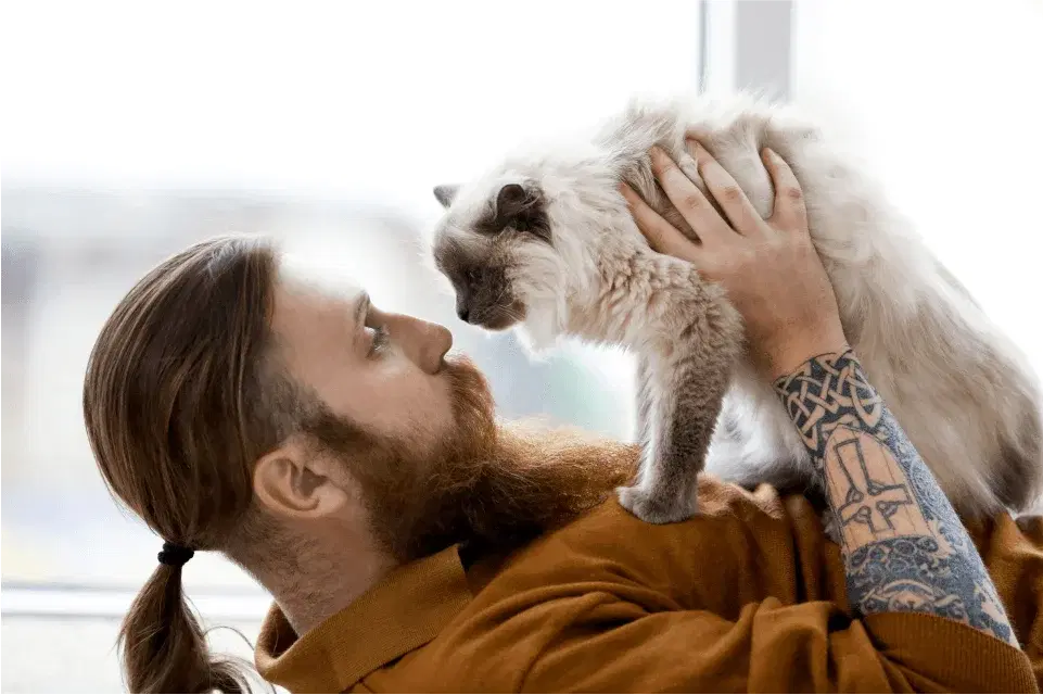 A bearded person with tattoos holds a fluffy cat close to their face.