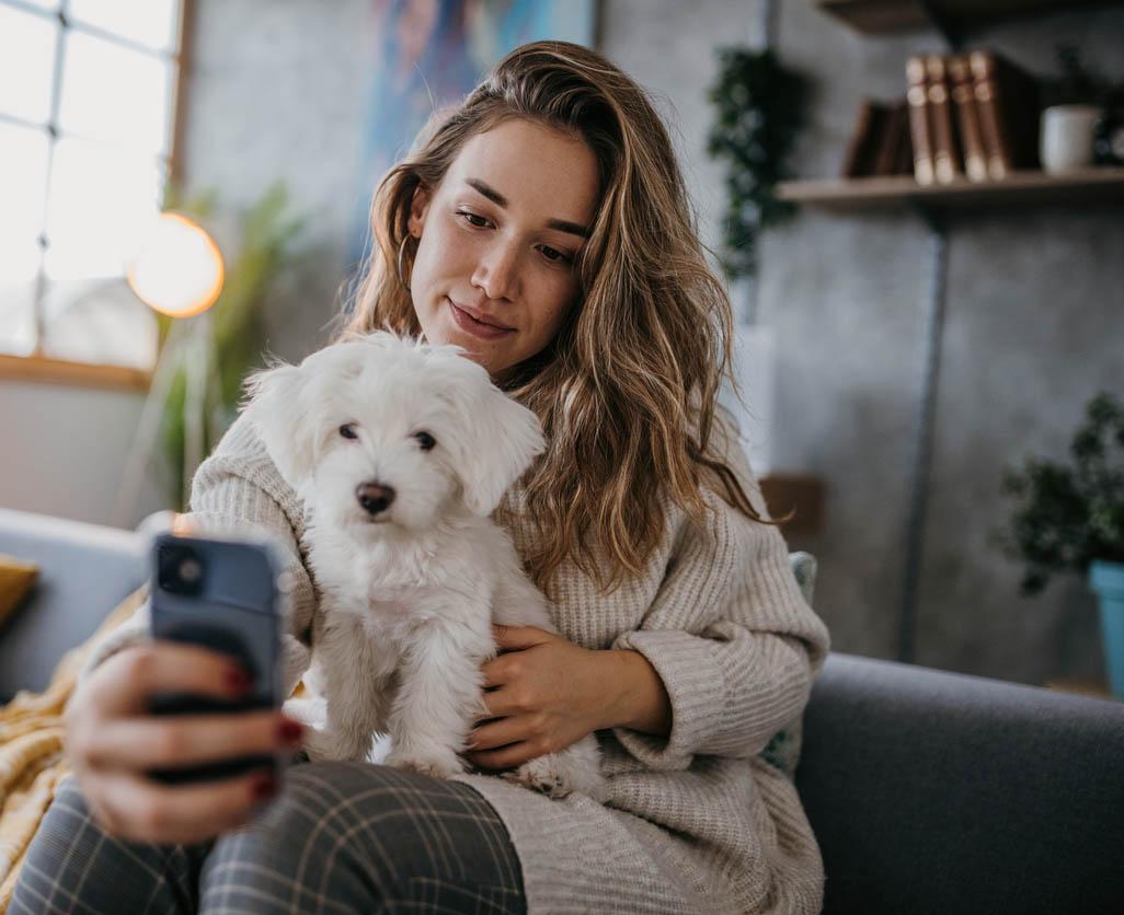 A woman with long hair holds a small white dog and takes a selfie on her phone.