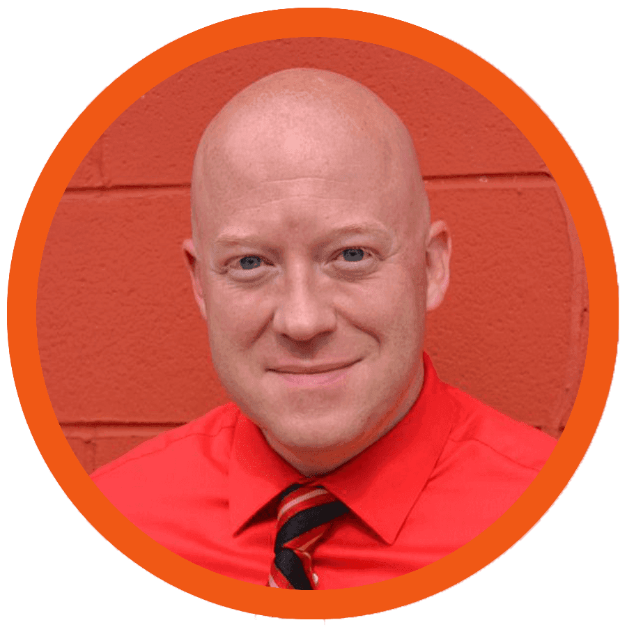 A bald man smiles against a red brick wall.